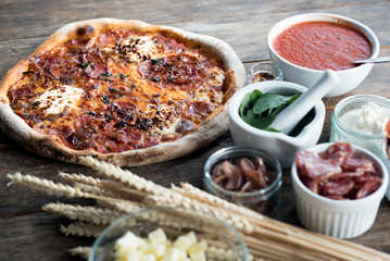 An amazing fresh pizza is laying on a wooden table with all the ingredients around, with the dried chilli rolled out as a heart form 