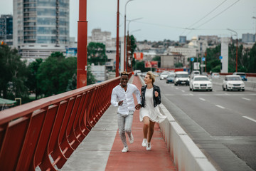 Naklejka premium Young interracial couple runs on bridge and laughs cheerfully on the background of the city and the road.