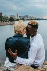 Interracial couple sits on rocks and hugs against background of river and city.
