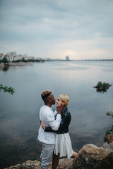 Interracial couple stands and hugs against background of river and city.