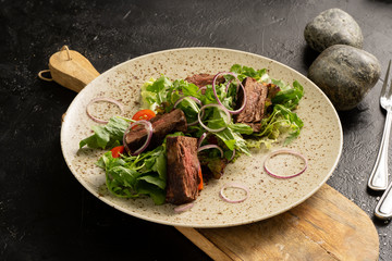 Beef roast beef with arugula, Beijing cabbage salad, onions, cherry tomatoes and chilli peppers. Plate with fried meat and salad on a black table.