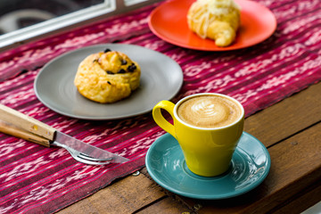 A cup of latte is served with colorful ceramic cups, side by side with food and cutlery. Everything is photographed on the table with traditional woven cloth ornaments ..