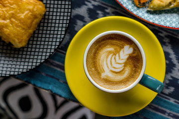 A cup of latte is served with colorful ceramic cups, side by side with food and cutlery. Everything is photographed on the table with traditional woven cloth ornaments ..