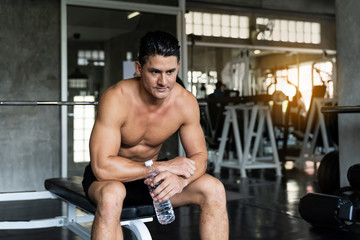 Fototapeta na wymiar Handsome muscular man sitting and drinking water after his workout in the fitness gym