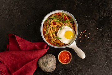 Noodles with beef, onions, eggs, vegetables and spicy adjika sauce. Plate with hot meat dish on a black table. View from above.