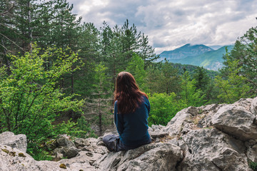 Naklejka premium A full body shot of an unrecognizable young Caucasian female hiker sitting on a rock in the French Alps mountains on a cloudy summer day