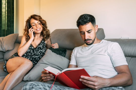 Young Couple Hanging Out At Home And Sitting On The Couch. The Boy With The Black Beard Reads A Red Book And The Girl Talks To The Family On The Phone.