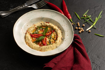 Homemade chickpea hummus with red chili pepper, onion and sunflower seeds on a black table.