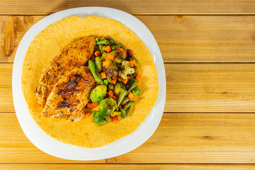 Grilled chicken breast and a mixture of vegetables on a corn tortilla in a white plate with copy space on a wooden background. Close-up