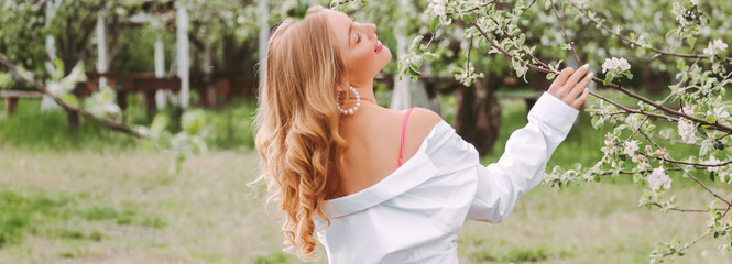 Panoramic garden young beautiful blonde woman with wavy hair in white shirt posing with blossom tree in green park. Cheerful cute carefree hippie girl relax and enjoy nature on countryside garden