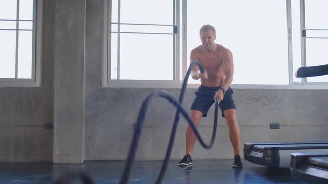 Slow motion of sport muscular man exercise in gym with battle rope with hot weather and day light. Concept of hard workout in gym to build healthy body for prevent disease and covid infection.