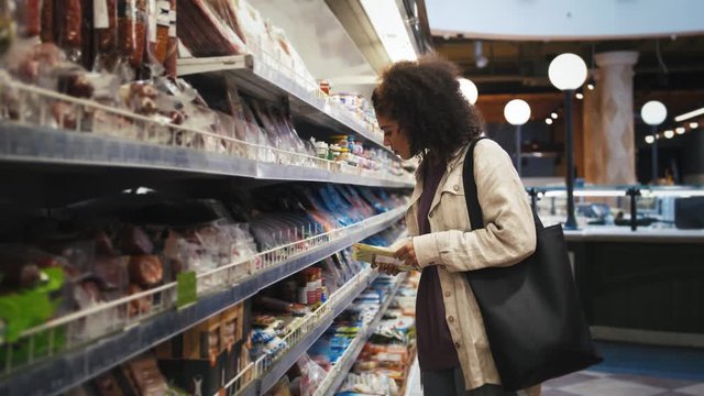 Dark-skinned girl in headphones and casual outfit. She is listening to the music and choosing sausages while shopping in a grocery