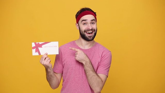 Young bearded fun fitness sporty guy 20s sportsman in headband pink t-shirt spend weekend in home gym hold gift coupon isolated on yellow background studio. Workout sport motivation lifestyle concept