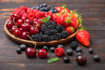 Fresh organic summer berries mix in round wooden tray on dark wooden table background. Raspberries, strawberries, blueberries, blackberries and cherries.