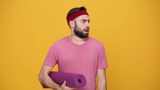 Young bearded fun fitness sporty guy 20s sportsman in pink t-shirt holds rolled up mat karemat crying take off headband isolated on yellow background studio. Workout sport motivation lifestyle concept