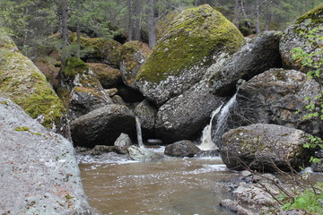 Naklejka na ściany i meble A mountain river waterfall flows through a river bed through huge boulders with moss
