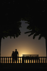 Silhouette of a couple in love on a sunset background. Palm trees and a bench. Sea view point. Vacation and tourism concept, travel. The relationship. Vertical photo. Happy together. Italy.