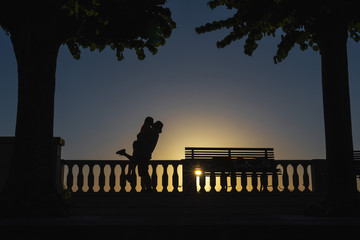 Silhouette of a kissing couple in love on a sunset background. Palm trees and a bench. Sea view point. Vacation and tourism concept, travel. The relationship. Happy together. Italy.