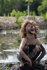 A young boy is playing in the mud, he is very dirty, messy and having a great time. The boy is laughing, happy, the kid feels freedom and free. He is outside, free range child. He is at summer camp