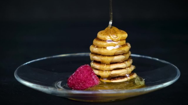 Tiny pancake cereal - trendy quarantine food. Slow motion honey pours on pyramid of mini pancakes in plate with fresh raspberry on black background.