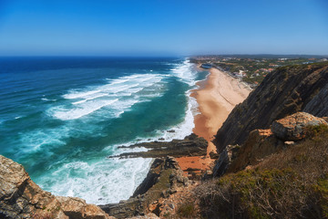 Rocky shore of the Atlantic Ocean. View of high cliffs, foamy waves and sandy beach. Sunny summer seascape. Portuguese riviera. In the area of Cabo da Roca. Portugal.