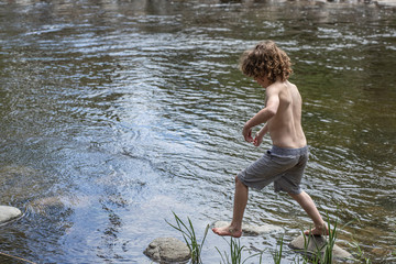A young boy without his shirt is jumping over rocks in a pond. He is playing in the water, in the summer at camp. Leaping across stones in the creek, pond, lake. Summer vacation, camp. Fun in sun