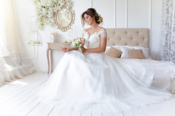 Fototapeta na wymiar A gorgeous bride in a beautiful dress is sitting on a bed in a bright room and holding a bouquet of flowers . Wedding. Wedding bouquet. morning of the bride. A happy bride is waiting for the groom.
