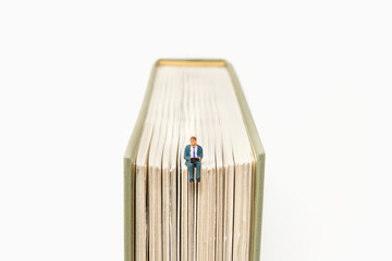 A businessman working on a laptop sits on top of a hardback book - Tiny People Working from Home...