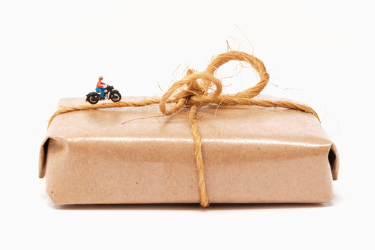 A motorcyclist rides over a classic isolated brown paper wrapped parcel tied with brown parcel string in a bow- Tiny People Working from Home Series