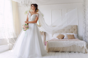 A gorgeous young bride in a white wedding dress in the room. A beautiful model stood at full height at the large window of the house. The girl is waiting for her fiance in a bright room.Wedding.