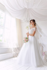 Fototapeta premium A gorgeous young bride in a white wedding dress in the room. A beautiful model stood at full height at the large window of the house. The girl is waiting for her fiance in a bright room.Wedding.