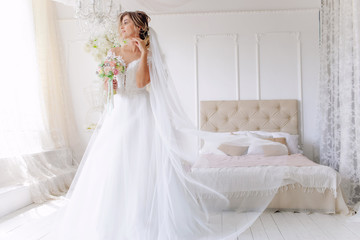Fototapeta na wymiar A gorgeous young bride in a white wedding dress in the room. A beautiful model stood at full height at the large window of the house. The girl is waiting for her fiance in a bright room.Morning 