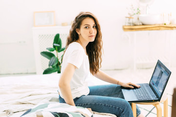 Young curly brunette on the bed in the bedroom with a laptop. Home office of a freelancer. Young...