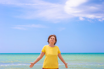 A cheerful elderly woman over 65 stands on the shores of the azure sea against a blue sky, spreads her arms out to the sides and enjoys a summer day