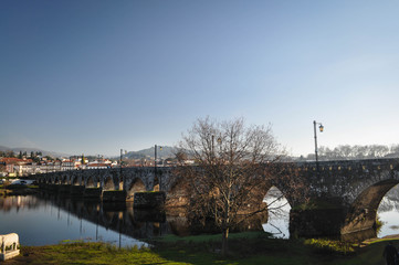 The old stone arch bridge across the river. City on the second side of the river. Tree without leaves in the foreground. Portugal.