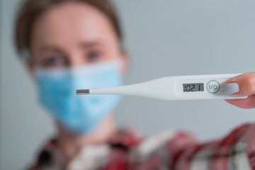 Woman in medical face mask showing digital medical thermometer with high temperature - selective focus, close up. Healthcare, measurement, disease, covid 19, infection, coronavirus concept