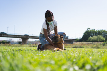 African woman with a mask on her face and braids caressing a boxer dog lying on the grass