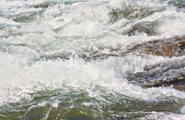 Obraz na płótnie Canvas Rapid spring river flowing over rocks on sunny day, forming white water waves, closeup detail - abstract nature background