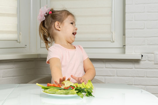 a little girl sits at a table in the kitchen and pushes away a plate of vegetables. children do not want to eat vegetables
