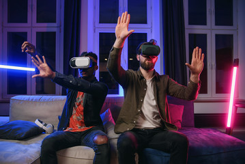 Fototapeta premium Portrait of positive likable satisifed multiracial male mates which sitting together on the couch in somebody's room and competing between each other in video game using special 3d goggles
