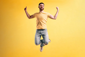 Fototapeta na wymiar Life is beautiful! Young man dressed in a yellow t-shirt and light jeans jumping against a yellow wall shows thumbs up and smiling.