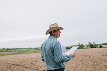 Man farmer agronomist in jeans and shirt stands back in field after haymaking, with tablet looking into the distance. Rural business, agricultural industry, freedom after work, concept