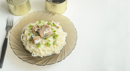 A glass plate with spaghetti and sliced green onions and an open tin of canned fish on a white background.The fork is on the table..Copyspace.