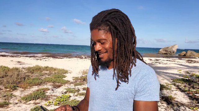 Millennial deadlock Rasta man smiles and laughs at international text message or social media while roaming in nature background at tropical beach in the Caribbean island of Barbados