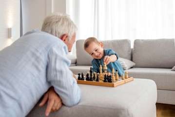 Smiling little boy playing chess with his grandfather at home