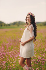 Pregnant happy girl stand and hold hands on stomach, stand on grass in the outdoor in the field of pink flowwers background with. Close up.