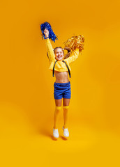 cute cheerleader girl in a yellow tank and blue shorts holds pompons and dances / jumps