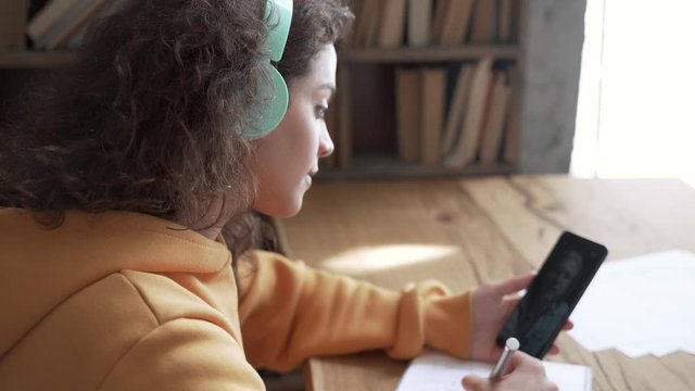Teen girl school college student wear headphones hold mobile phone distance learning online class math lesson with teacher tutor using zoom app video conference call write notes. Over shoulder view.