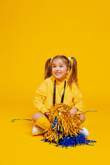 cute cheerleader girl in a yellow tank top and blue shorts sits with pompons in her hands
