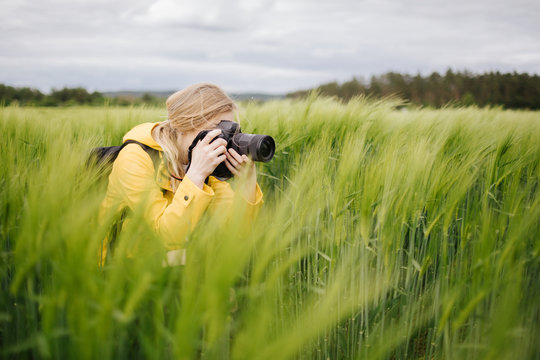 Charming blonde photographer in yellow jacket taking pictures of green wheat field. Mature lady using professional digital camera for creating amazing photos on fresh air.